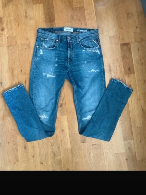 Blå slitna replay jeans - Säljer ett par blå jeans från replay med slitningar och distressed detaljer framtill och på bakfickorna. Klassisk femficksmodell i rak passform och tillverkade i denim. Perfekta för en avslappnad och trendig look. Storlek 30 midja och 34 längd.