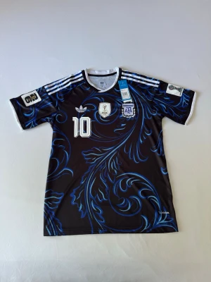 Argentina Messi tröja svart/blå Adidas - Snygg svart Argentina fotbollströja från Adidas med blått mönster, vita detaljer och nummer 10. Messi och landslagets emblem på bröstet. Korta ärmar, klassiska Adidas-ränder på axlarna och tryck på ryggen. Perfekt för fotbollsfans!