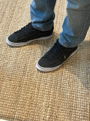 Svarta Polo Ralph Lauren sneakers - Snygga svarta sneakers från Polo Ralph Lauren med klassisk låg profil och vit sula. Skorna har svart ovandel i textil med diskret logga i guld på sidan och svarta skosnören. Perfekta för en clean och stilren look.