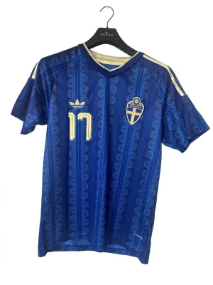 Blå Sverige fotbollströja Adidas - Säljer en blå Sverige fotbollströja från Adidas med nummer 17 och namnet Gyökeres på ryggen. Tröjan har gula detaljer, korta ärmar och ett mönster i tyget. Perfekt för dig som vill stötta landslaget på match eller träning.