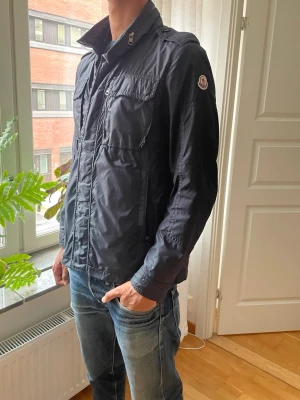 Blå Jacka från Moncler - Snygg svart vindjacka från Moncler med hög krage, bröstfickor med lock och dragkedja framtill. Jackan har en klassisk Moncler-logga på ärmen och justerbar krage med en huva i som går att ta ut. Perfekt för dig som vill ha en stilren och funktionell jacka både till svala sommarkvällar, hösten och våren.  Jackan har dock en liten defekt se sista bilden, men det är inget man ser med blåtaögat vid användning så de påverkat ej den som bär jackan😃