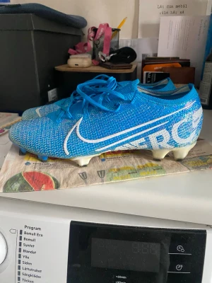 Nike Mercurial Vapor 13 Elite FG blå - Säljer ett par Nike Mercurial Vapor 13 Elite FG fotbollsskor i blått med vita detaljer och stor Nike-logga på sidan. Skorna har snörning, låg profil och är tillverkade i ett lätt syntetmaterial för bästa grepp på naturgräs.