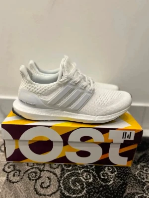 Vita Adidas Ultra Boost sneakers - Säljer ett par helvita Adidas Ultra Boost sneakers med stickad ovandel i mesh och klassiska tre ränder på sidan. Skorna har en bekväm Boost-sula och vita snören. Perfekta för dig som gillar stilrena och sportiga sneakers.