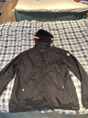 Svart vindjacka från Moncler - Snygg svart vindjacka från Moncler med huva och dragkedja framtill. Jackan har två fickor med knappar och en Moncler-logga på ärmen. Huvan har en röd och vit detaljkant. Perfekt för blåsiga dagar. Priset är inte hugget i sten, går att diskutera
