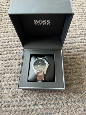 Silverfärgad klocka från Hugo Boss - Stilren klocka från Hugo Boss med svart urtavla och silverfärgat armband i metall. Klockan levereras i originalask med tydlig logga. Perfekt accessoar för dig som gillar klassisk och modern design.