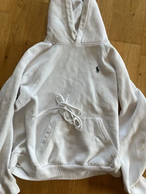 Vit hoodie från Polo Ralph Lauren - Klassisk vit hoodie från Polo Ralph Lauren med broderad marinblå logga på bröstet. Hoodien har huva med dragsko, känguruficka framtill och ribbade muddar. Tillverkad i mjuk bomullsblandning som känns skön mot huden. Perfekt för en avslappnad och stilren look. Band till luvan medföljer 