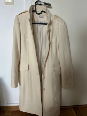 Krämvit ullkappa med axelklaffar - Stilren krämvit kappa från Filippa K i ullblandning. Kappan har axelklaffar, knäppning framtill med stora knappar och två fickor med lock. Insidan är fodrad med randigt tyg. Perfekt för dig som gillar klassisk och clean stil.