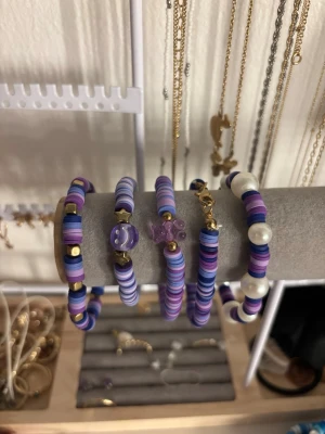 Lila och vita pärlarmband set - Säljer ett set med fem armband i lila, vitt och blått. Armbanden är gjorda av platta pärlor i olika nyanser, vissa med gulddetaljer och en med vita pärlor. Ett armband har en lila blomformad pärla och ett annat har en lila pärla med prickar. Perfekt för dig som gillar färgglada accessoarer.