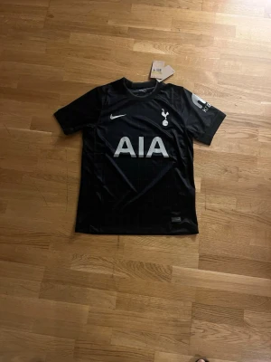 Tottenham Hotspur svart matchtröja Nike - Snygg svart Tottenham Hotspur matchtröja från Nike med diskret rutmönster, klubbmärke och Nike-logga på bröstet samt AIA-tryck framtill. Tröjan har rund hals och korta ärmar, perfekt för fotboll eller träning.  Fraktar dagen eller samma dag som du köper.