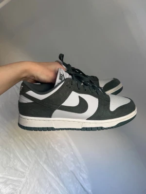 Nike Dunk Low svart/vit sneakers - Snygga Nike Dunk Low sneakers i svart och vitt med klassisk swoosh på sidan. Skorna har lågt skaft, perforerad tåbox och ovandel i mocka och läder. Perfekta för dig som gillar streetwear och vill ha en stilren look.