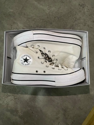 Vita Converse Chuck Taylor Platform - Säljer ett par vita Converse Chuck Taylor All Star med hög platåsula och klassisk svart rand. Skorna är höga och har canvasmaterial, vita skosnören och metallfärgade öljetter. Ikonisk Converse-logga på sidan. Perfekta för dig som vill ha en clean och tidlös look. Flexibel med priset