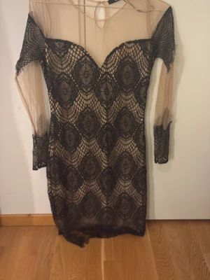 Svart spetsklänning från Bik Bok - Snygg kort klänning från Bik Bok med svart spetsmönster över beige tyg. Klänningen har långärmade meshärmar och en v-ringad urringning. Perfekt för dig som vill sticka ut med en unik och elegant look.