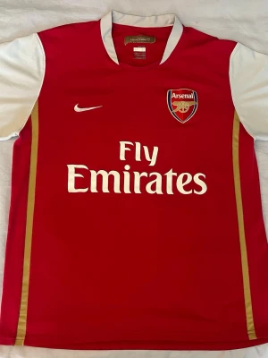 Arsenal Vintage Retro T-Shirt HENRY 14 - Röd Arsenal matchtröja från Nike med vita ärmar och gulddetaljer. Tryck med Fly Emirates på bröstet och Arsenal-logga. På ryggen står det HENRY och nummer 14. Perfekt för fotbollsfans och samlare.