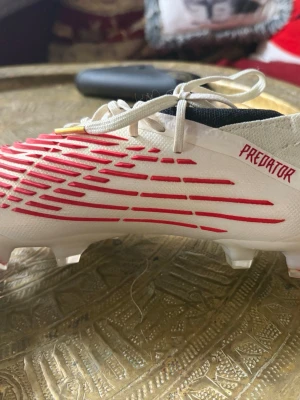 Adidas Predator vita fotbollsskor - Säljer ett par vita Adidas Predator fotbollsskor med röda detaljer och texten 'Predator' på sidan. Skorna har snörning, plastdobbar och är tillverkade i syntetmaterial för bra grepp och kontroll på planen. Perfekta för dig som vill sticka ut på matchen.