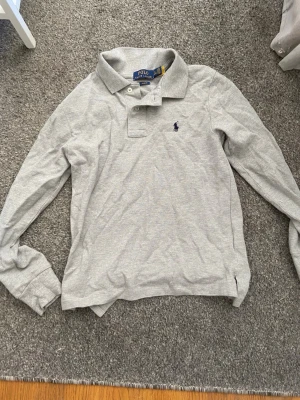 Grå långärmad piké från Polo Ralph Lauren - Klassisk grå långärmad pikétröja från Polo Ralph Lauren med blå broderad logga på bröstet. Slim fit-modell med krage och knappar framtill. Tillverkad i mjuk bomull som känns skön mot huden.