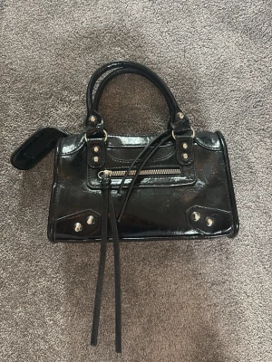 Svart city bag - Säljer en svart city bag  med silverfärgade detaljer, dragkedja framtill och dubbla handtag. Väskan har dekorativa nitar och långa läderband som ger en cool vibe. Perfekt för dig som gillar statement-accessoarer.