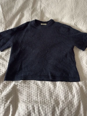 Mörkblå stickad t-shirt - En enkel och stilren mörkblå stickad t-shirt med korta ärmar och rund hals. Tröjan har en rak passform och ribbstickade kanter vid ärmslut och nederkant. Perfekt för dig som gillar minimalistisk stil.