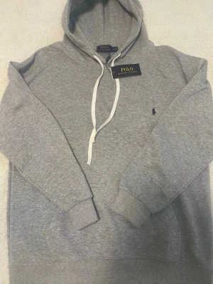 Grå hoodie från Polo Ralph Lauren - Snygg grå hoodie från Polo Ralph Lauren med klassisk huva och vita dragsnören. Tröjan har ribbade muddar och en liten mörkblå broderad logga på bröstet. Tillverkad i mjukt bomullsmaterial som känns skönt mot huden. Perfekt för en avslappnad och stilren look.