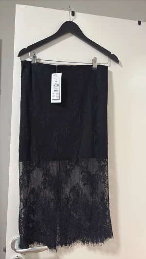 Svart lång spetskjol - Snygg svart långkjol från Gina Tricot med transparent spets och blommigt mönster. Kjolen har en rak passform och vacker volangkant nedtill. Perfekt för dig som vill ha en trendig och elegant look.