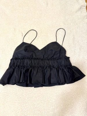 Svart volang topp med smala band - Snygg svart topp med smala axelband och volangdetaljer. Toppen har en rynkad midja som ger en cool och trendig look. Perfekt att styla med jeans eller kjol för en chic outfit.