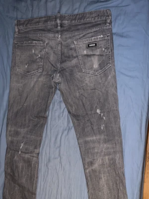 Grå slim jeans från Dsquared2 - Säljer ett par grå slim fit jeans från Dsquared2 med slitna detaljer och klassisk femficksmodell. Jeansen har en snygg tvättad look och Dsquared2-logga på bakfickan. Tillverkade i Italien i bomullsmix. Perfekta för dig som gillar streetwear och trendig stil.