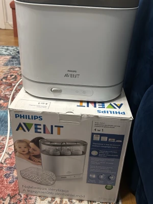 Philips Avent flasksterilisator - Elektrisk flasksterilisator från Philips Avent i vitt och grått. Rymmer flera nappflaskor samtidigt och har en modern, fyrkantig design. Enkel att använda med tydliga knappar på framsidan. Perfekt för att hålla flaskor och tillbehör rena.