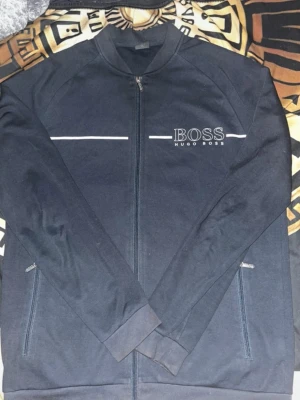 Svart ziptröja från Hugo Boss - Snygg svart ziptröja från Hugo Boss med vit logga och diskreta vita detaljer på bröstet. Tröjan har hög krage, dragkedja framtill och två fickor med dragkedja. Perfekt för en clean och stilren look.