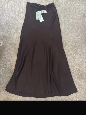 Svart långkjol i satin - Snygg svart långkjol i mjukt satinliknande material med lätt glans. Kjolen har en enkel och stilren design med en diskret söm som ger en cool detalj. Perfekt att matcha med en crop top eller stickad tröja.