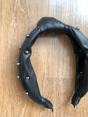 Svart diadem med silvriga nitar - Snyggt svart diadem i fejkläder med puffig form och coola silvriga nitar längs hela bandet. Perfekt accessoar för att ge din look edge och attityd. Materialet är mjukt och diademet sitter bekvämt på huvudet.