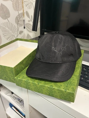 GG keps nylon  - Snygg svart keps från GG med diskret GG-monogram över hela kepsen. Framsidan är i glansigt tyg och baksidan har meshpanel för extra ventilation. Klassisk böjd skärm och justerbar rem bak. Perfekt för dig som gillar exklusiva accessoarer. Box medföljer 