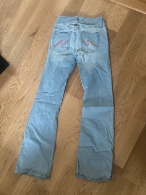 Ljusblå bootcut jeans med rosa söm - Säljer ett par ljusblå bootcut jeans med rosa zigzag-detaljer på bakfickorna. Jeansen har hög midja och klassisk femficksdesign. Materialet är mjukt denim i bomull och passformen är avslappnad med utsvängda ben.