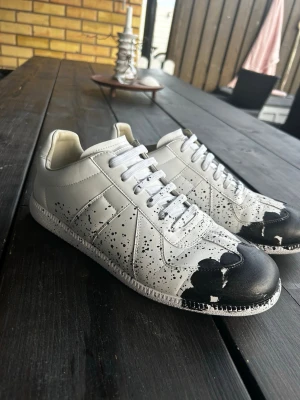Maison Margiela svartvit sneakers - Unika sneakers från Maison Margiela i vitt läder med svart färgstänk och svartmålade tår. Klassisk låg modell med vita skosnören och detaljerad sula. Perfekta för dig som gillar statement-plagg och streetstyle.