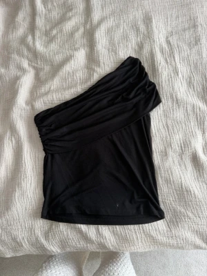 Svart oneshoulder topp från nelly  - Säljer en svart oneshoulder topp från nelly. Toppen har en asymmetrisk axel och är tillverkad i mjukt stretchigt material som sitter snyggt mot kroppen. Perfekt för dig som vill ha en stilren och trendig look.