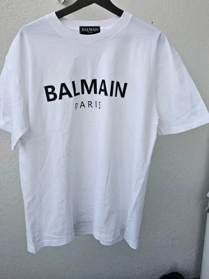 Vit Balmain t-shirt med tryck - Vit t-shirt från Balmain med svart logotyp-tryck på bröstet. Klassisk rund halsringning och korta ärmar. Tillverkad i mjuk bomull för en skön och stilren look.