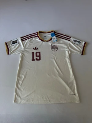 Helt ny Spanien fotbollströja Adidas Lamine Yamal #19 - Säljer en vit Spanien fotbollströja från Adidas med vinröda detaljer och nummer 19. Tröjan har Lamine Yamal tryckt på ryggen, officiellt landslagsmärke på bröstet och klassiska Adidas-ränder på axlarna. Tillverkad i lätt och funktionellt material, perfekt för match eller träning.