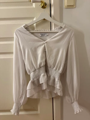 Vit blus med smock och volanger - Supersöt vit blus med smock i midjan och vid ärmslut. Blusen har långa ärmar, v-ringning och fina volangdetaljer nedtill. Osäker vad original priset är 