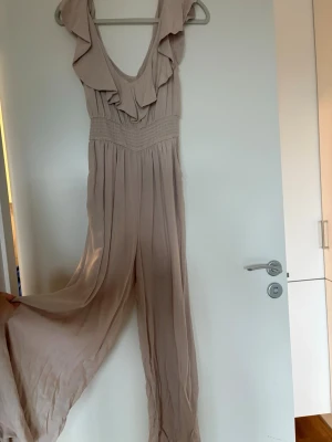 Beige jumpsuit med volangdetaljer - Säljer en beige jumpsuit med vida ben och volangdetaljer vid axlar och urringning. Djup rygg och smock i midjan för skön passform. 