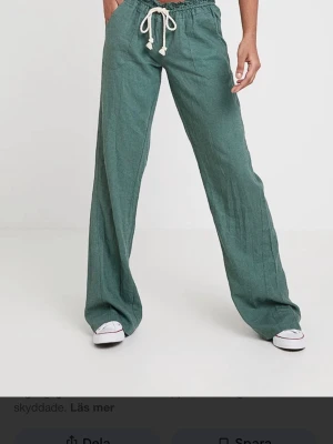 Gröna Roxy linnebyxor  - Snygga gröna linnebyxor med avslappnad passform och breda ben. Byxorna har resår i midjan och vit dragsko för skön och justerbar passform. Perfekta för en chill och trendig look. Två stora fickor framtill.