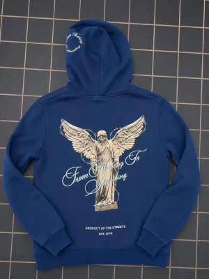 Blå hoodie från Hoodrich med ängeltryck - Snygg blå hoodie från Hoodrich med stort ängeltryck och texten 'From Nothing To Something' på ryggen. Framsidan har mindre tryck med logga och samma motiv. Huva med broderad text och klassisk känguruficka. Perfekt för en streetwear-look.