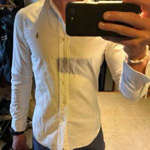 Vit skjorta från Ralph Lauren - Klassisk vit skjorta från Ralph Lauren med broderad logga på bröstet. Skjortan har lång ärm, knappar framtill och en snygg krage. Perfekt för dig som gillar stilrena och tidlösa plagg.