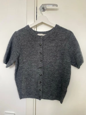 Grå stickad kortärmad kofta - Mysig grå stickad kofta med korta ärmar och rund halsringning. Koftan har knappar framtill och är gjord i ett mjukt, ull-liknande material. Perfekt att slänga över en topp för en avslappnad look.