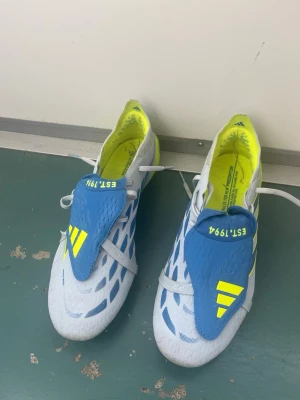 Adidas Predator fotbollsskor blå/gul elite - Säljer ett par Adidas Predator fotbollsskor i vitt, blått och neon-gult. Skorna har snörning, mönstrad ovandel och texten 'EST. 1994' på plösen. Sulan är gul med blå detaljer och grepp för konstgräs eller gräs. Perfekta för dig som vill sticka ut på planen!