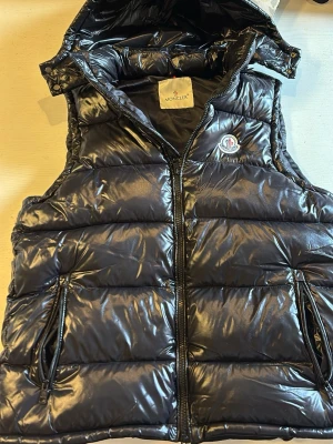 Svart dunväst från Moncler - Snygg svart dunväst från Moncler med glansig finish och avtagbar huva. Västen har två dragkedjeförsedda fickor och klassisk Moncler-logga på bröstet. Insidan har ett unikt serietidningsmönster och märkestaggar. Perfekt för lager-på-lager-stil. Västen här iprincip helt ny, har bara testat på den och säljer den pga att den inte passade mig. Pris kan diskuteras lite. Hör av er vid frågor eller funderingar 😇