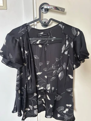 Svart blommig omlottblus Zara Woman - Svart omlottblus från Zara Woman med vita blommönster. Blusen har korta, fladdriga ärmar och ett lätt, luftigt tyg som ger en avslappnad känsla. Perfekt till jeans eller kjol för en trendig look. Står m men skulle säga s