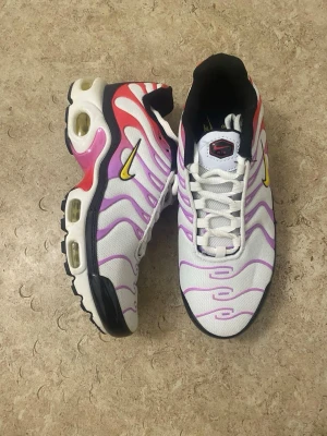 Nike Air Max Plus vita/lila sneakers - Nike Air Max Plus sneakers i vitt mesh med lila och rosa vågiga detaljer, gula swoosh-loggor och svarta samt röda inslag. Synlig Air Max-dämpning i sulan och svart yttersula. Ikonisk streetstil med sportig vibe och bekväm passform.