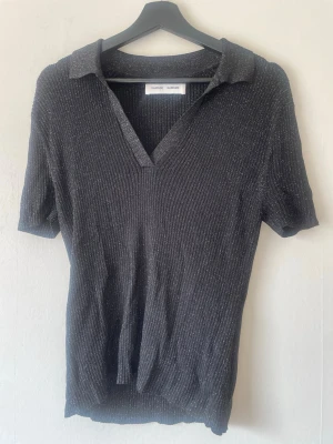 Glittrig svart topp från Samsøe Samsøe - Snygg svart glittrig topp från Samsøe Samsøe med ribbad struktur och v-ringning.  modell med krage, perfekt för dig som vill ha något extra till din outfit. Materialet känns mjukt och stretchigt.