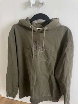 Olivgrön hoodie från GANT - Snygg olivgrön hoodie från GANT med broderad logga och texten 'American Sportswear' på bröstet. Klassisk huva med dragsko och ribbade muddar vid ärmar och nederkant. Perfekt för en avslappnad och trendig look.