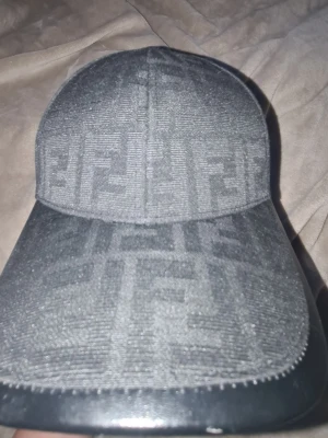 Svart keps med Fendi-logga - Snygg svart keps från Fendi med det klassiska FF-monogrammet över hela kepsen. Kepsen har böjd skärm och är tillverkad i textil med detaljer i läderimitation på skärmens kant. Perfekt accessoar för en trendig streetstyle-look.