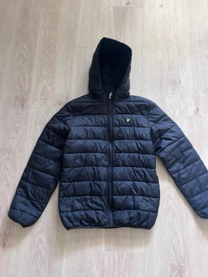 Svart pufferjacka från Lyle & Scott - Snygg svart pufferjacka från Lyle & Scott med huva och dragkedja framtill. Jackan har det klassiska Lyle & Scott-logotypen i gult på bröstet och quiltad design som ger en sportig look. Perfekt för kalla vinterdagar.