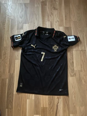 Portugal Ronaldo matchtröja svart - Snygg svart Portugal matchtröja från Puma med guldiga detaljer och nummer 7. Ronaldo står på ryggen och tröjan har officiella märken på ärmarna. Tillverkad i lätt och ventilerande material, perfekt för fotboll eller träning.